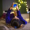 A girl in a long-sleeved blue shirt is lying on her bed, holding her phone.  Her bedroom is decorated with strings of white lights.