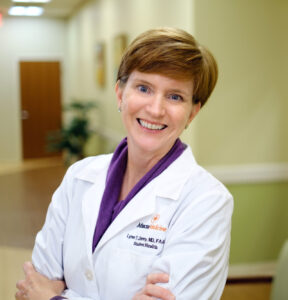 Dr. Lynn Denny is wearing a white lab coat and is standing with her arms crossed in the office hallway.