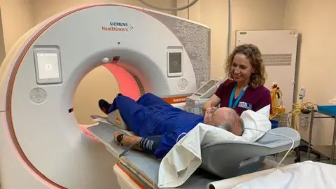 BBC Man is lying down trying to get into the CT scanner with a nurse next to him